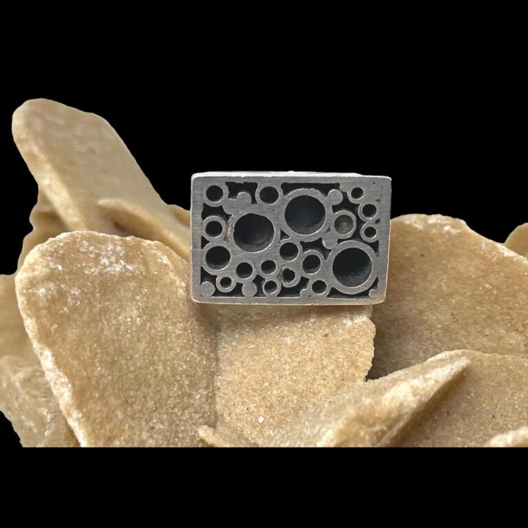Main photo of Modernist Brutalist silver ring displayed on top of a rock of natural desert sand rose. The ring front is the only visible part of the ring as it is quite a substantial size. The silver is unpolished and gives an impression it is metal rather than sterling silver. There are bubble like crater clusters on the ring 3 larger than the rest. Very unique and fascinating ring!