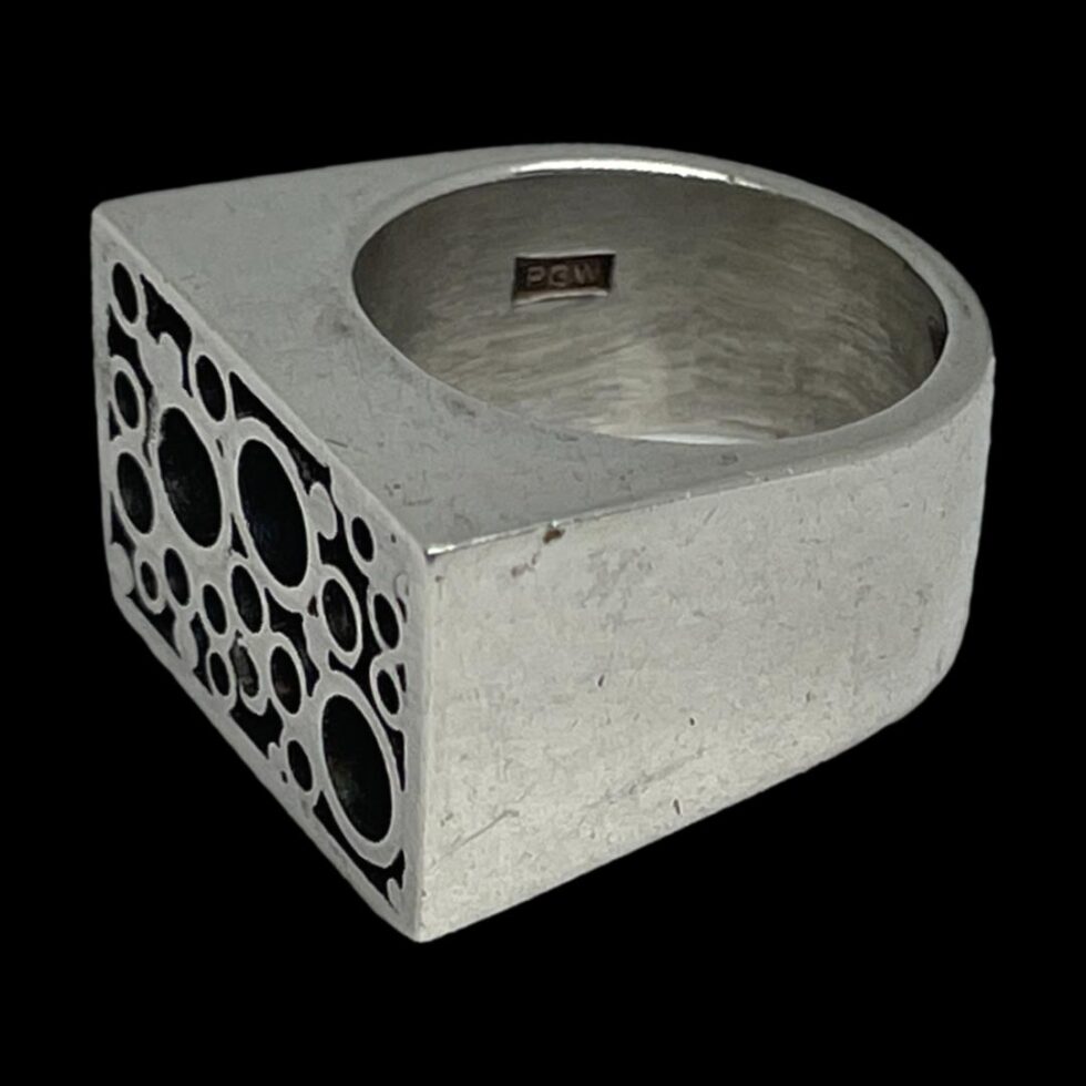 Photo of ring displayed on a flat surface with a black background this time with the ring front facing towards the bottom left corner. The PGW initials for Peter Guy Watson is visible to the thick inside band.