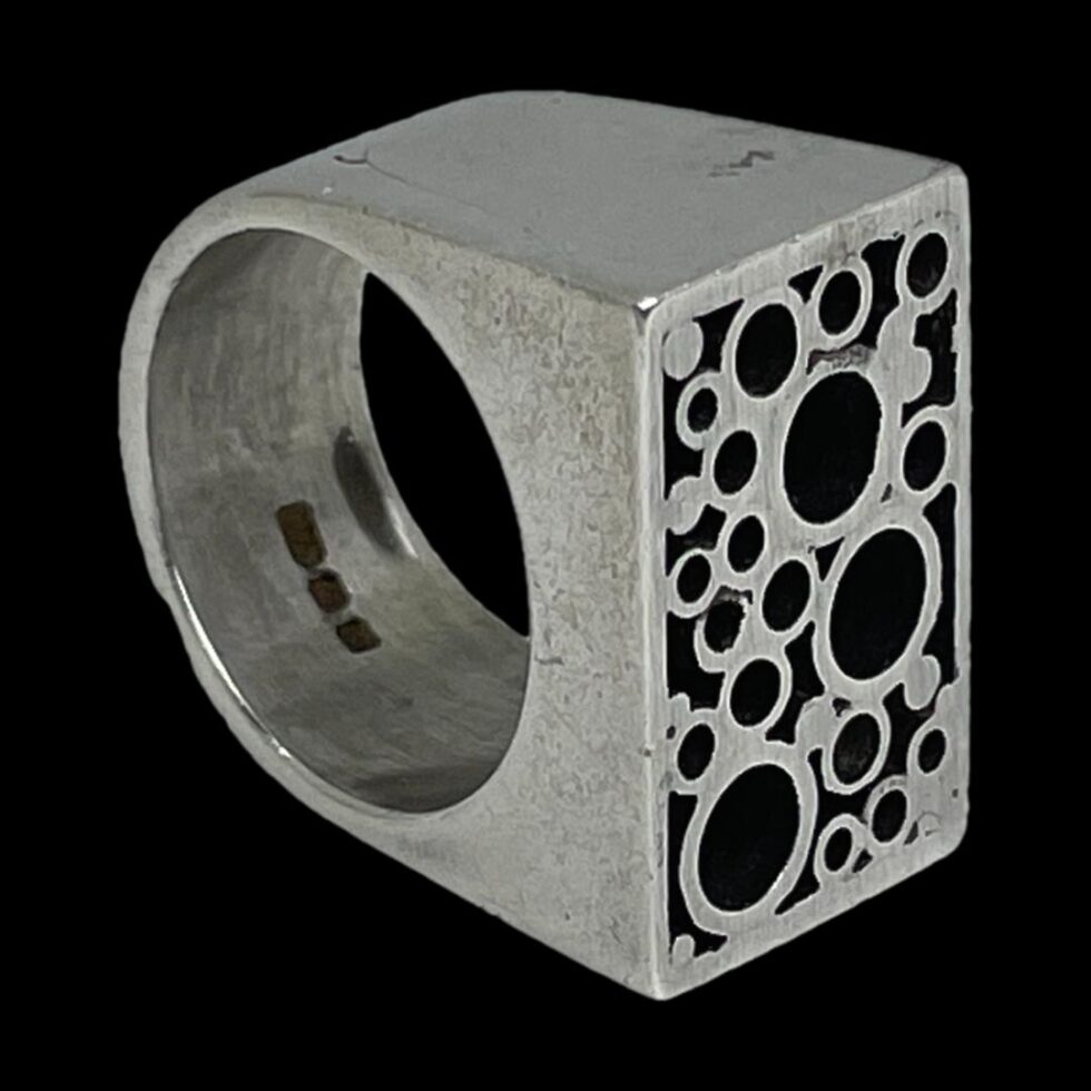 Photo of Modernist Brutalist ring displayed on its side with the ring front to the bottom right corner. The hallmark is visible to the inside band (quite difficult to make out)