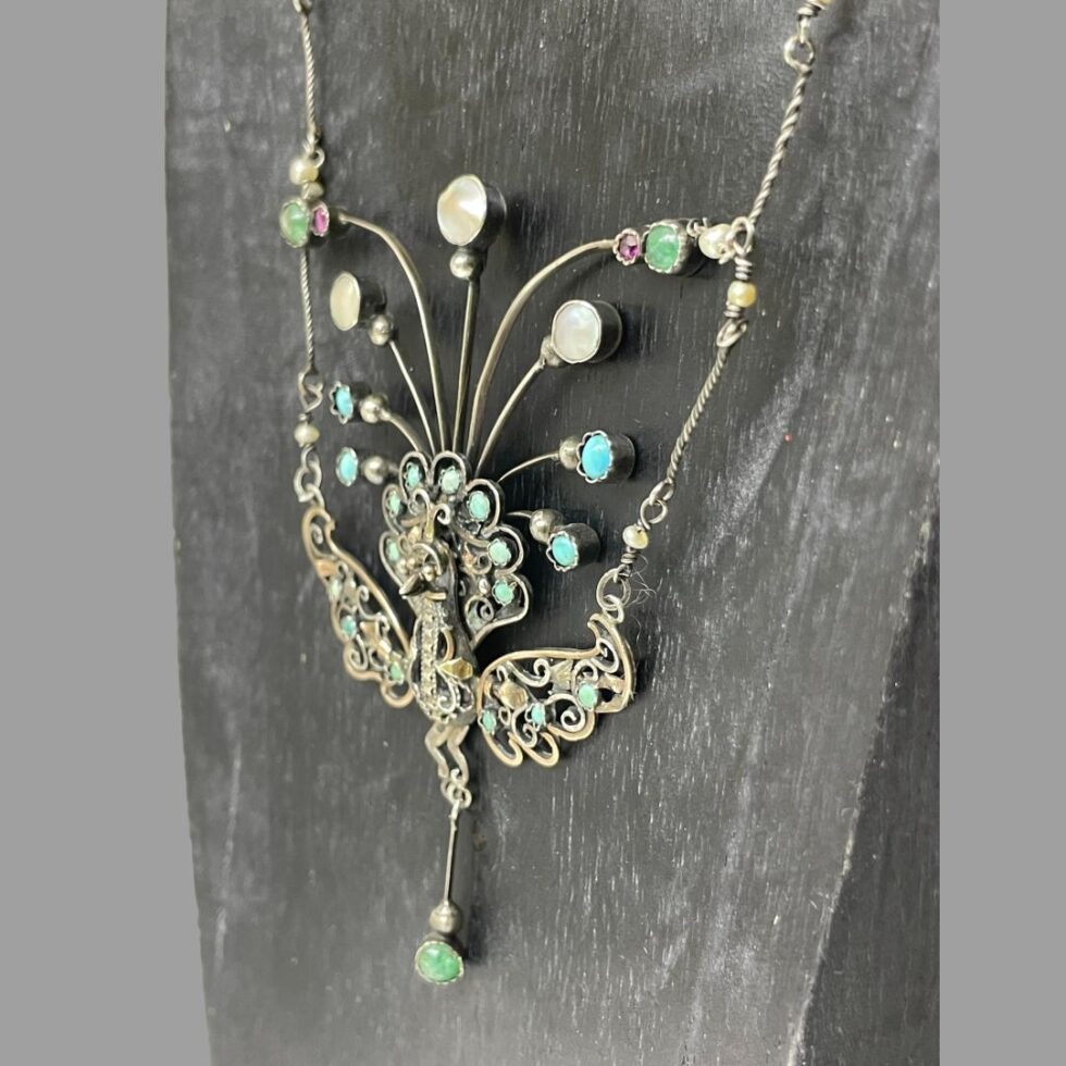 Photo of the peacock area of necklace still displayed on a black wooden stand this time with the front facing to the bottom left corner.
