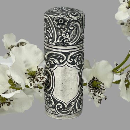 Main photo of silver portable perfume bottle displayed from a near eye level with lid shut and the cartouche in the foreground. There is a spray of white orchid laid behind it for display. The empty cartouche is shaped like a shield and the rest of the bottle has floral engravings throughout.