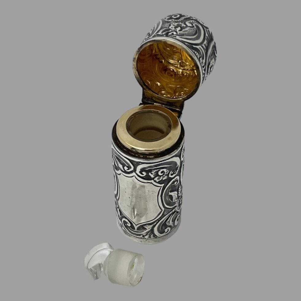 Photo of silver portable perfume bottle displayed from a slightly raised angle looking diagonally down. The lid is fully open on its hinge revealing the gilt interior and the glass stopper has been removed and placed just in front of the cartouche area which is facing to the bottom left corner of photo.