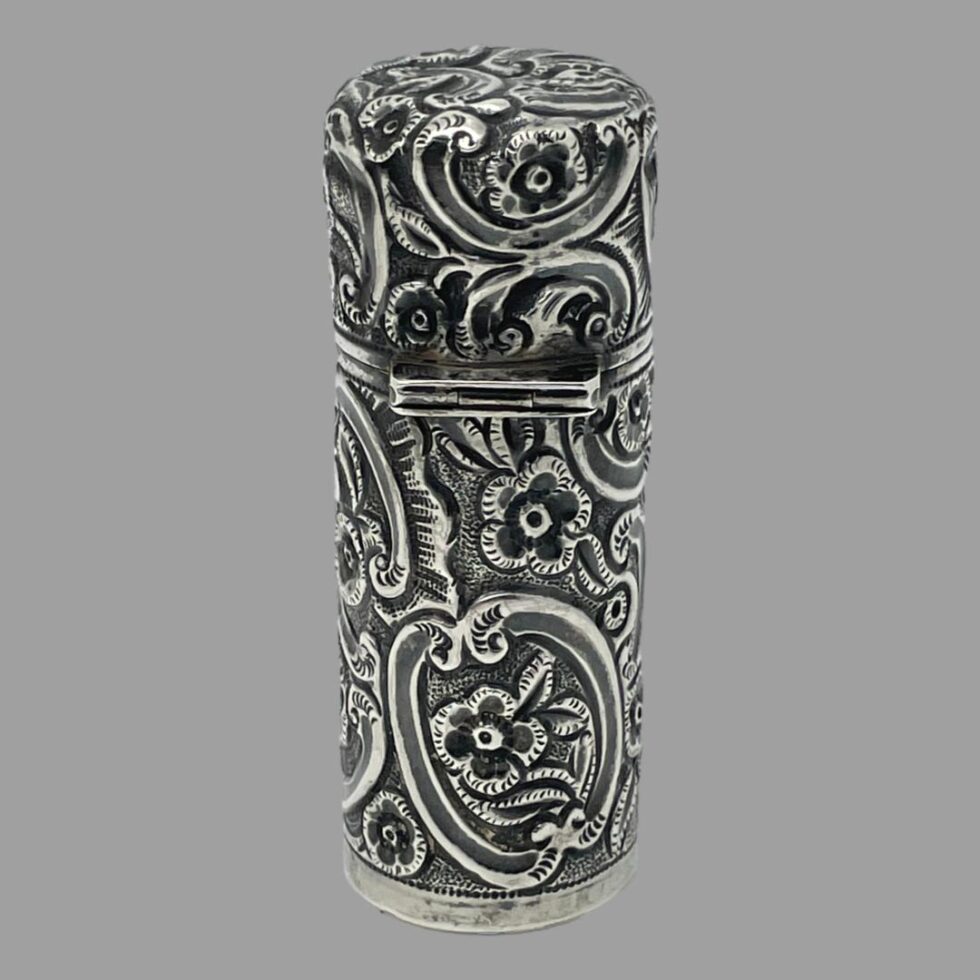 Photo of silver portable perfume bottle displayed upright, closed and from the back showing the intricately engraved floral design detail which covers the whole item except for the cartouche area.