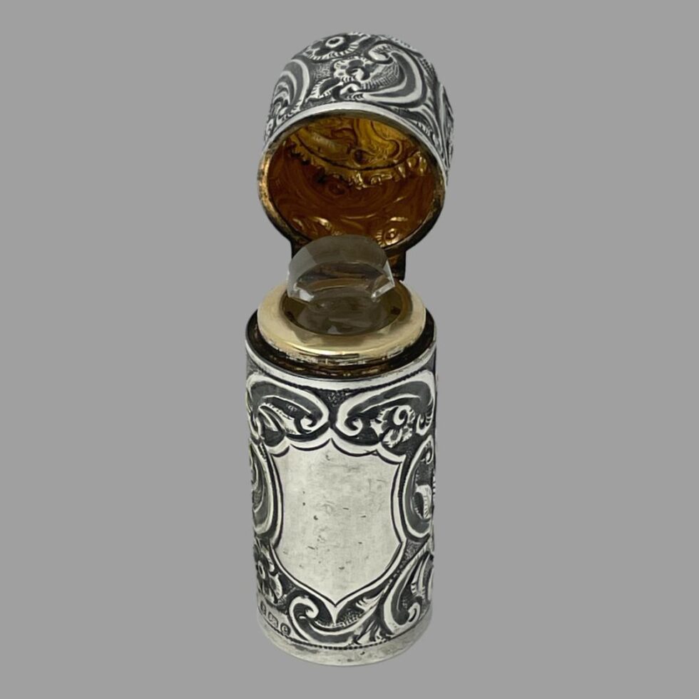 Photo of silver portable perfume bottle displayed upright with top hinged open and with the cartouche in the foreground. The glass stopper for the bottle is in its place.