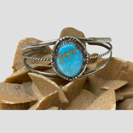 Main photo of Native American turquoise cuff displayed on top of a rock of natural desert sand rose and shown with the front forward facing. The turquoise is a beautiful shade and has some natural golden coloured inclusions. The silver frame around the turquoise stone is a good thickness with some simple yet effective debossed line designs. The cuff has 3 rows of silver bands ; 2 plain at top and bottom and a twisted one at the centre. These gather to a point at the end of the cuff and smoothed off.