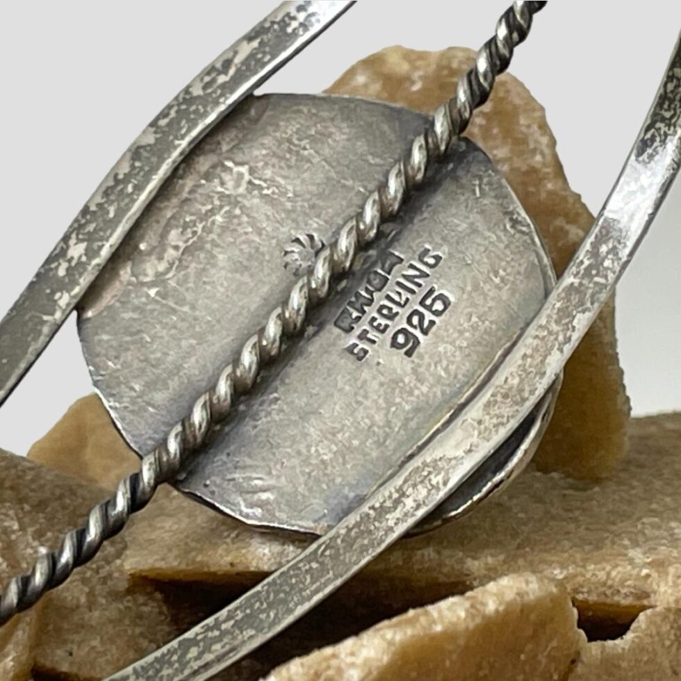 Close up photo of the back of the Native American turquoise cuff showing the hallmark and initials of the maker. The back of the stone setting is closed and the three bands attached.