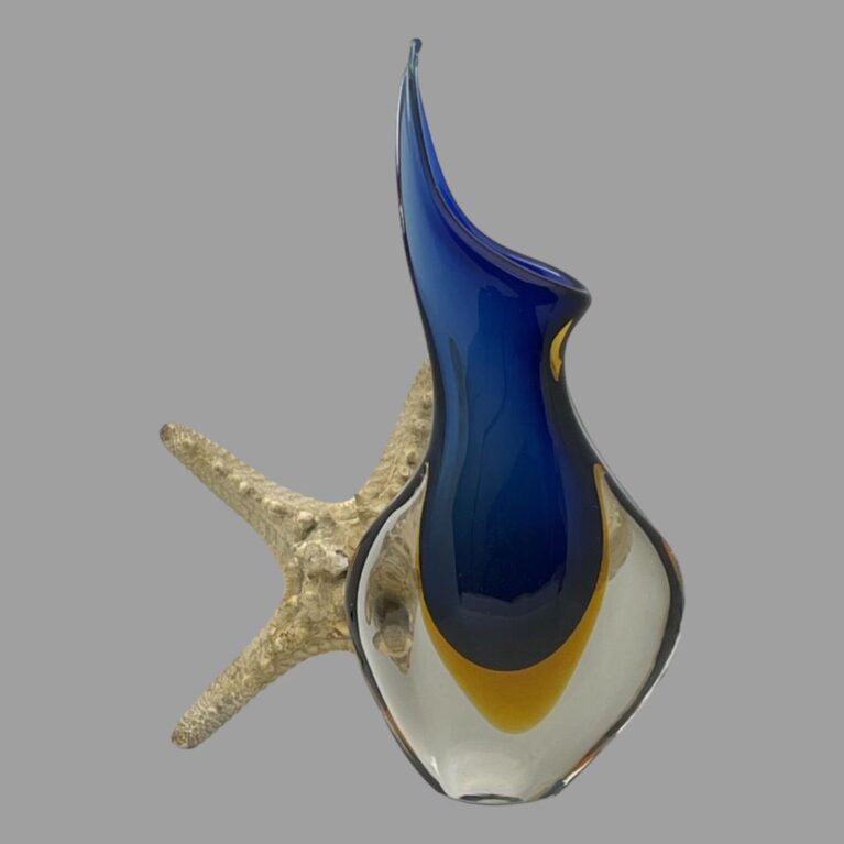 Main photo of Murano blue sommerso vase seen from the wider flatter angle. Like a lot of vases, this one has a wide side and a narrow side. Here we are looking at the wider side. The vase has an interesting form with one top side extended to a point upward and higher than the other side. The lower part of the vase balloons out to the side almost like hips. The lower 'hip' area is in clear glass with an egg yolk yellow centre in the middle. The rest of the vase is a beautiful rich blue and this starts from just above the egg yolk yellow all the way to the top. The clear glass also covers an area of the blue at the bottom. There is a spray of white orchids starfish placed behind and to the left of the vase for display only.