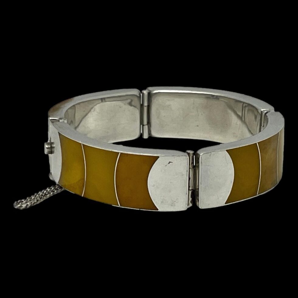 Photo of Modernist Silver amber bracelet displayed on a flat surface with black background. The clasp is shown closed to the left of photo with the safety chain protruding out. One of the hinged areas is in the foreground. This bracelet has 3 hinged areas plus the clasp.