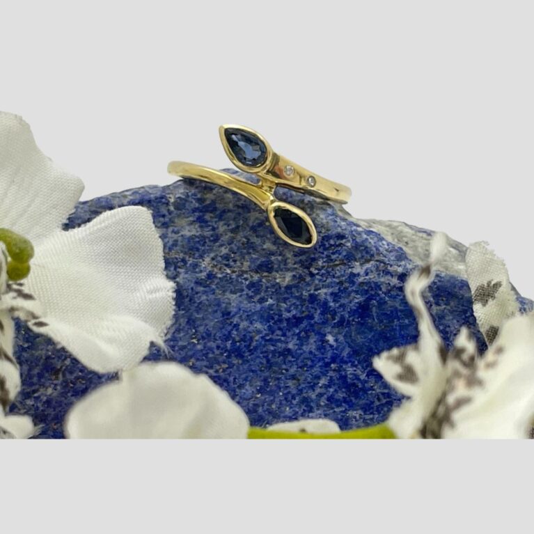 Main photo of gold sapphire diamond ring displayed on a small rock of natural lapis lazuli with the ring front forward facing. The ring has a split band at the front but it is a solid split with a piece of gold joining the ends solid just before the sapphires. In this photo, the pear cut sapphire is at the top with the 2 round cut diamonds visible to its right in the band. The drop cut sapphire below is to the right and below the round cut diamonds so when worn the ring has the sapphires diagonally across the finger. There are parts of white orchids around the lower part of the lapis stone.