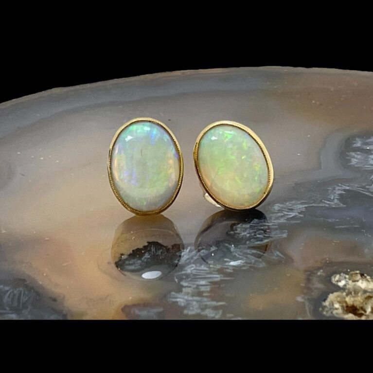 Main photo of gold &opal cabochon studs displayed on a slice of natural agate and seen with the earring front forward facing. The generous sized opals appear each framed in gold with the one on right leaning in slightly to the left. The colours within these opals are incredible.