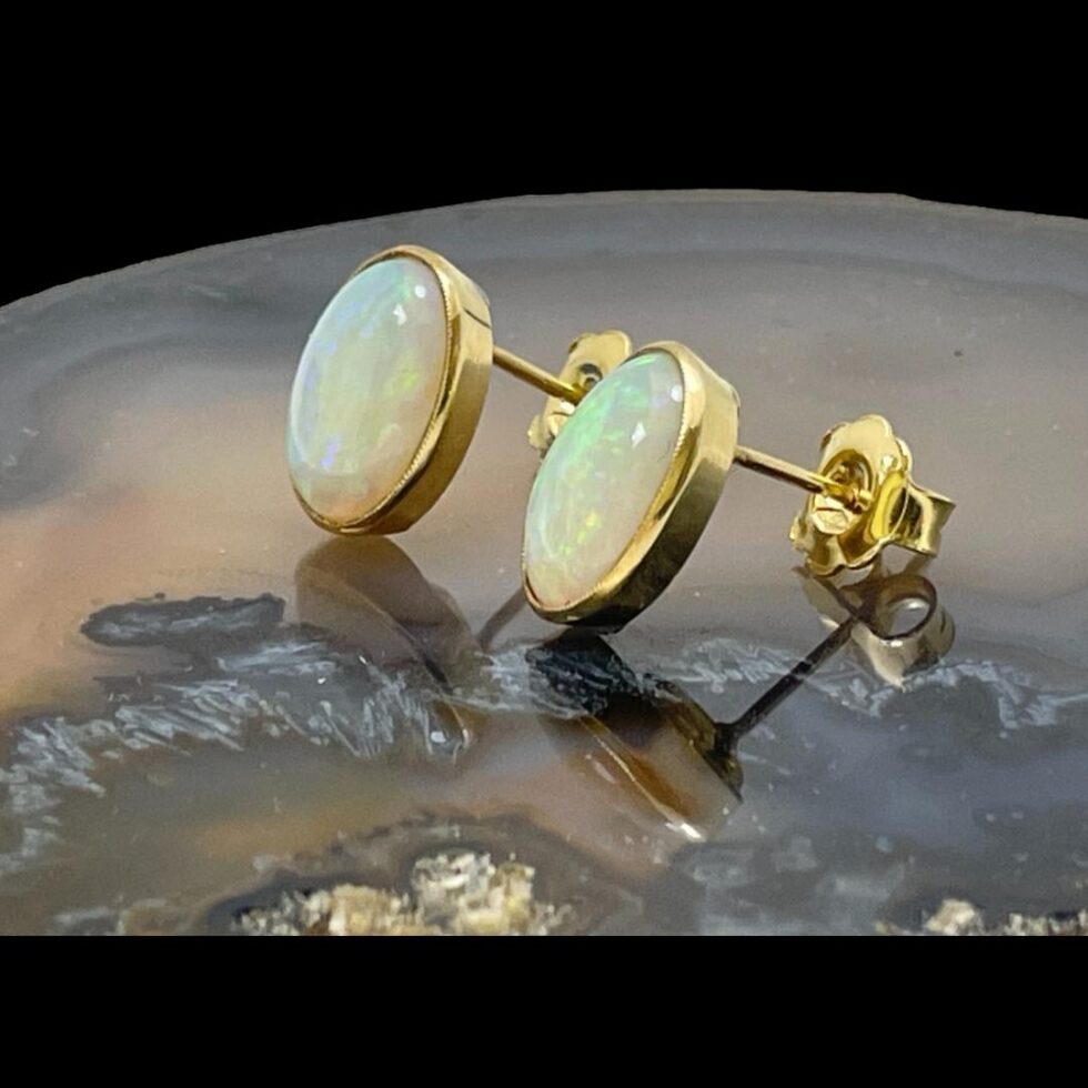 Photo of gold opal stud earrings still displayed on a slice of natural agate and seen with the earring fronts facing to the left. Each opal is well set in a generous width gold frame. The earrings are propped upright by their butterfly fasteners.