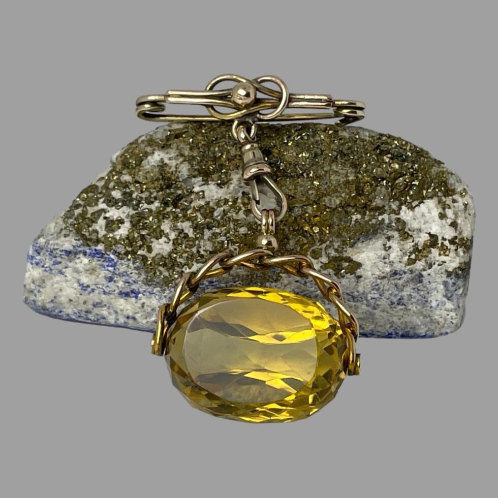 Main photo of antique gold citrine fob displayed with the faceted large citrine in the centre foreground and in front of a rock of natural lapis (hardly any blue showing) The pin and short chain is resting on the lapis rock. One of the 3 flat sides is in the foreground and the faceting detail is also visible all around the flat area. The citrine stone is held by a short rigid chain of just 7 links which is then attached by dog clasp to the pin which resembles a reef not design with a gold ball at the centre.