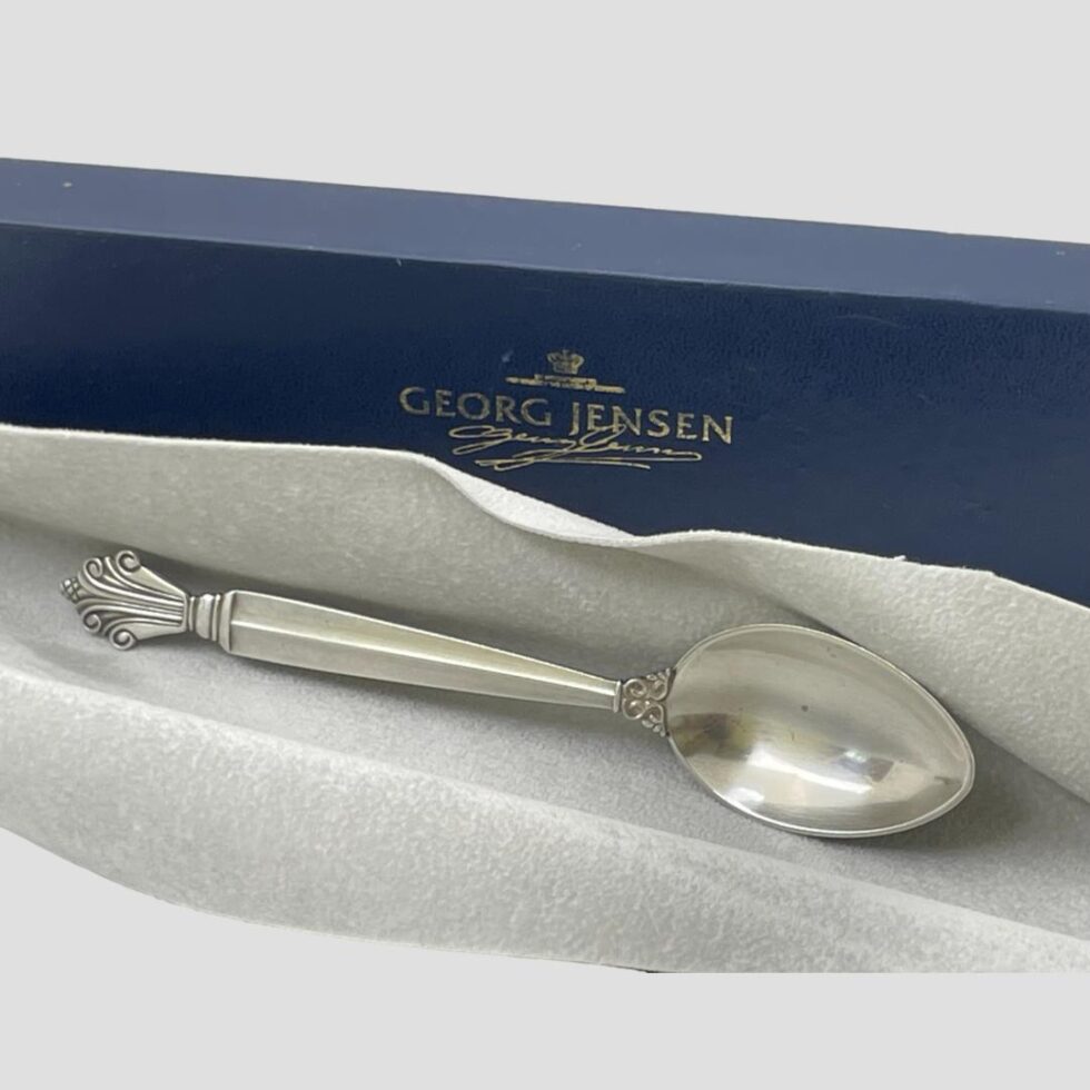 Photo of Jensen sterling silver spoon displayed in the box on top of its grey protector cloth. The spoon is propped on its side with the bowl on right and forward facing. The Georg Jensen box is behind.