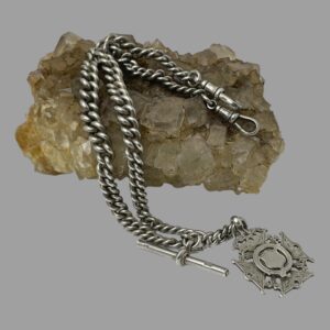 Main photo of antique sterling silver Albert watch chain displayed with fob and T-bar in the foreground on the flat and the chain and dog clips laid on top of a rock of river quartz. The chain graduates with the widest part in the middle of chain length.