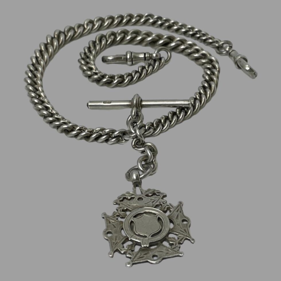 Photo of antique silver Albert chain displayed with the fob in the centre foreground , the chain which is laid out in a circular coil like a snake with one dog clip in the centre of coil and other on the outside to the middle right. Above the fob and part of chain is the T-bar