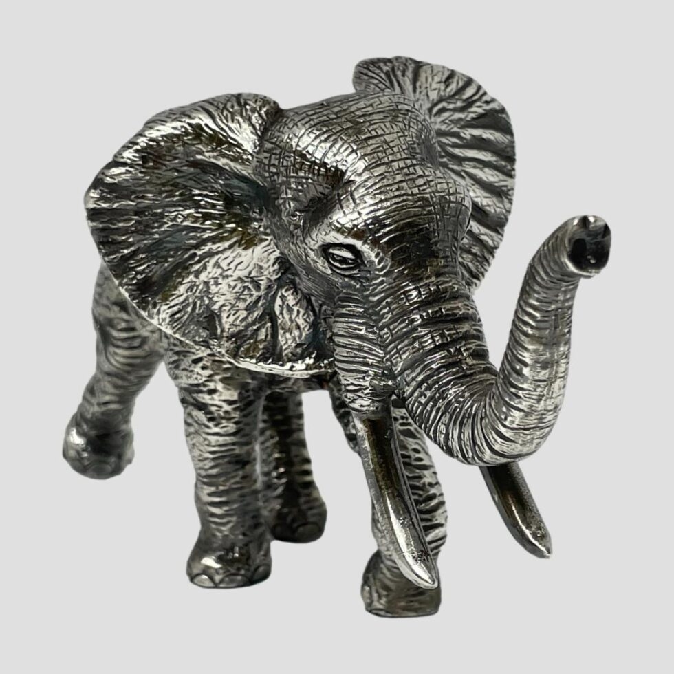 Photo of silver encased African elephant displayed facing the bottom right corner. Just like an African elephant, its head is huge with large ears. The trunk is raised and tusks are long. Great attention to detail.