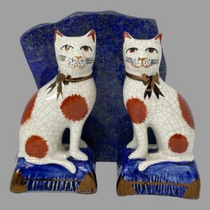 Main photo of Staffordshire Potteries seated cats posed in front of a rock of natural lapis lazuli and facing forward. The blue of the stone is almost identical to the blue of the cats cushions. The cats are off white in colour with dark terracotta spots and both have a golden brown ribbon around the neck.
