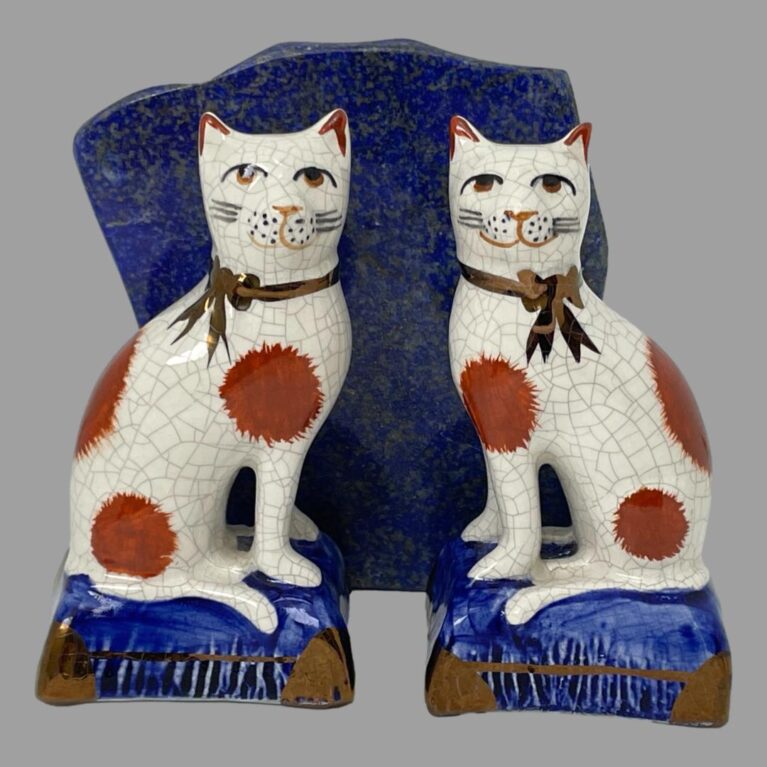 Main photo of Staffordshire Potteries seated cats posed in front of a rock of natural lapis lazuli and facing forward. The blue of the stone is almost identical to the blue of the cats cushions. The cats are off white in colour with dark terracotta spots and both have a golden brown ribbon around the neck.