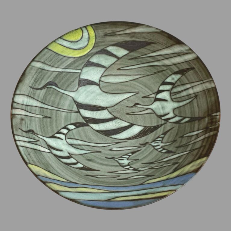 Main photo of large flying cranes bowl displayed looking straight into the decorated inside of bowl. The background base colour is of different shades of grey lines in a swirl matching the natural turn marks of the bowl. To the top left edge is a yellow sun with a band of white inside. Wispy white clouds are dotted about and 5 cranes are seen flying towards the left. The cranes are different in size giving depth with the largest in the foreground. They are white with black stripes to the wings. At the bottom is the sea in 2 shades of blue with yellow patches interpreted as the suns reflection on the water. The combination of grey lines in the background, wispy white clouds, black stripes on wings and the siege of cranes in flight gives this piece a mysterious movement - you feel as though you are flying with the birds. Wonderful piece of art.