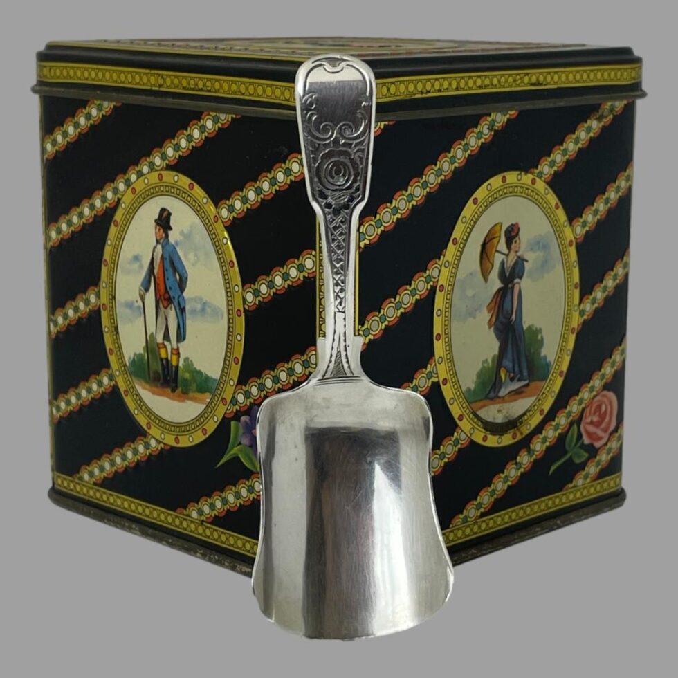 Photo of antique tea caddy spoon displayed leaning up against one corner edge of the tea caddy tin. The handle has some pretty scrollwork design with a stylised rose to the centre.