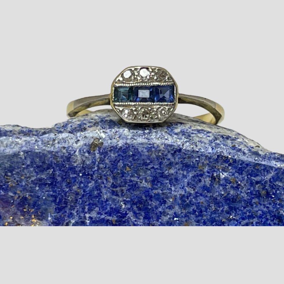 Main photo of Deco sapphire diamond ring displayed on a rock of natural lapis lazuli with the ring front forward facing. The stones are set in platinum with a row of square cut sapphires horizontally across the middle with the round cut diamonds in a row above and below. The ring front has an irregular hexagon shape which gives an extra Deco look.