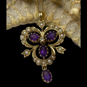 Main photo of gold pendant brooch &chain displayed leaning against a section of starfish (looks like seeded bread) and with no light shining. The amethysts are coloured a rich dark purple. The main body of the pendant or brooch is like an upside down club playing card suit with seed pearls all around the outer frame. At the centre, there is a single round cut diamond which then has 3 oval cut amethysts attached each housed within the space of the club. A single oval cut amethyst hangs below and 3 tulip shaped gold segments protrude from the dips of the club shape each with a seed pearl to the centre.