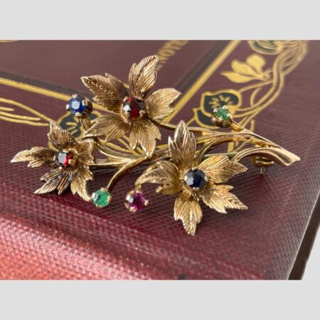 Main photo of gold &gemstones floral spray brooch displayed forward facing on the corner of a burgundy coloured old book. The brooch is laid across the bottom corner of the book cover which has a pretty gilt decoration. There are 3 floral blooms; top 2 with garnets to the centre and lower one with sapphire. There are 4 buds, 2 of emerald, 1 ruby and 1 sapphire. The flower petals and spray are in 9 karat gold. Lovely gift item for that special someone or yourself!