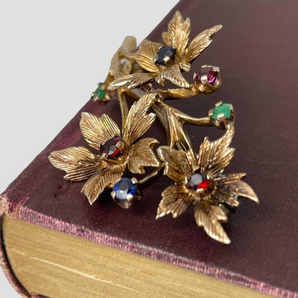 Photo of gold &gemstones brooch still displayed on a corner of an old book this time without gilt decoration. The brooch is displayed at an angle so the garnet blooms are in the foreground.