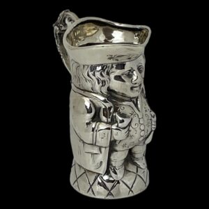 Main image of Edwardian silver Toby jug with front facing to the bottom right corner. The photo is taken from a little height showing a little of the hat interior. There is good attention to detail on this piece with different techniques used to create the caried textures for hair, waistcoat and base whilst the rest is smooth silver. The handle is quite elaborate with foliage like swirls and curls and a thumb rest on the top curve for a steady pour!
