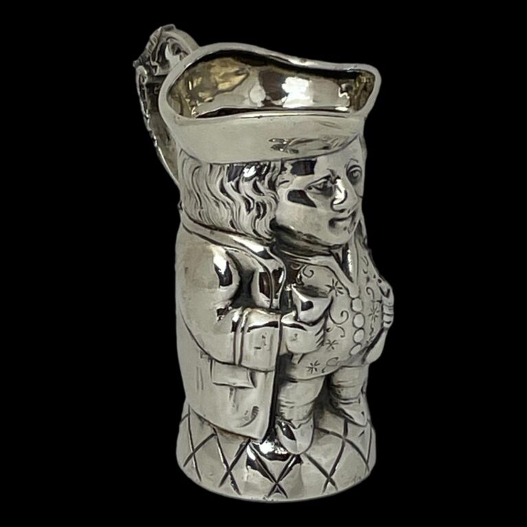 Main image of Edwardian silver Toby jug with front facing to the bottom right corner. The photo is taken from a little height showing a little of the hat interior. There is good attention to detail on this piece with different techniques used to create the caried textures for hair, waistcoat and base whilst the rest is smooth silver. The handle is quite elaborate with foliage like swirls and curls and a thumb rest on the top curve for a steady pour!