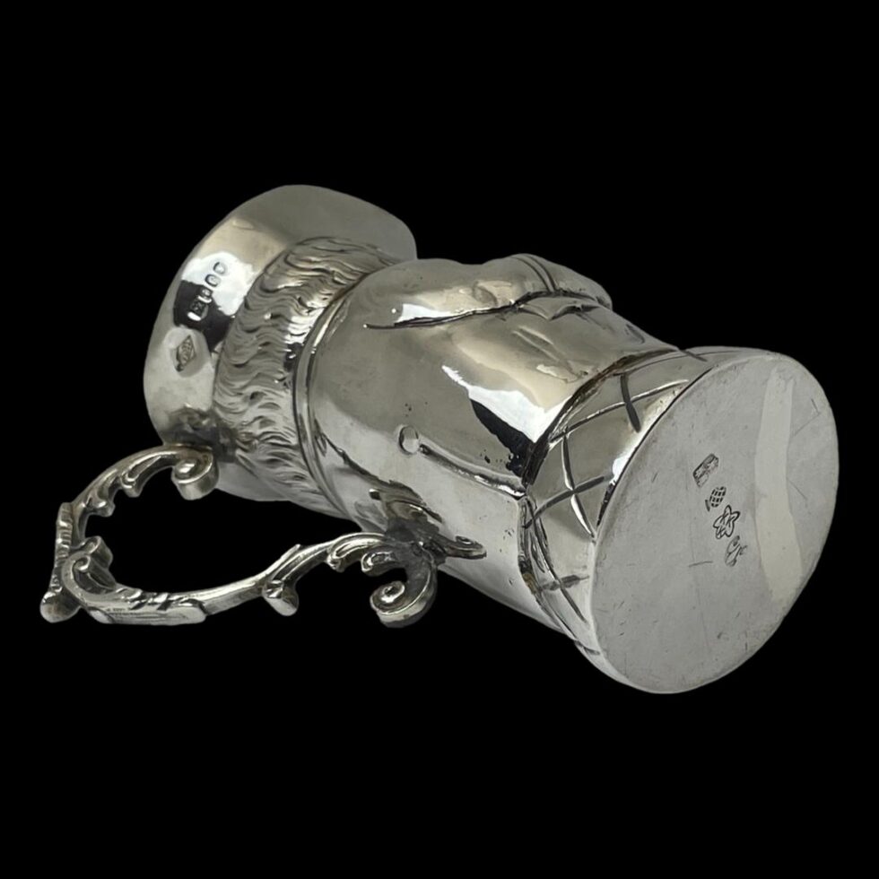 Photo of Edwardian silver Toby jug displayed laid flat on its side with the handle in the bottom left corner. The German hallmark is visible at the base and the British hallmark around the tricorn hat.