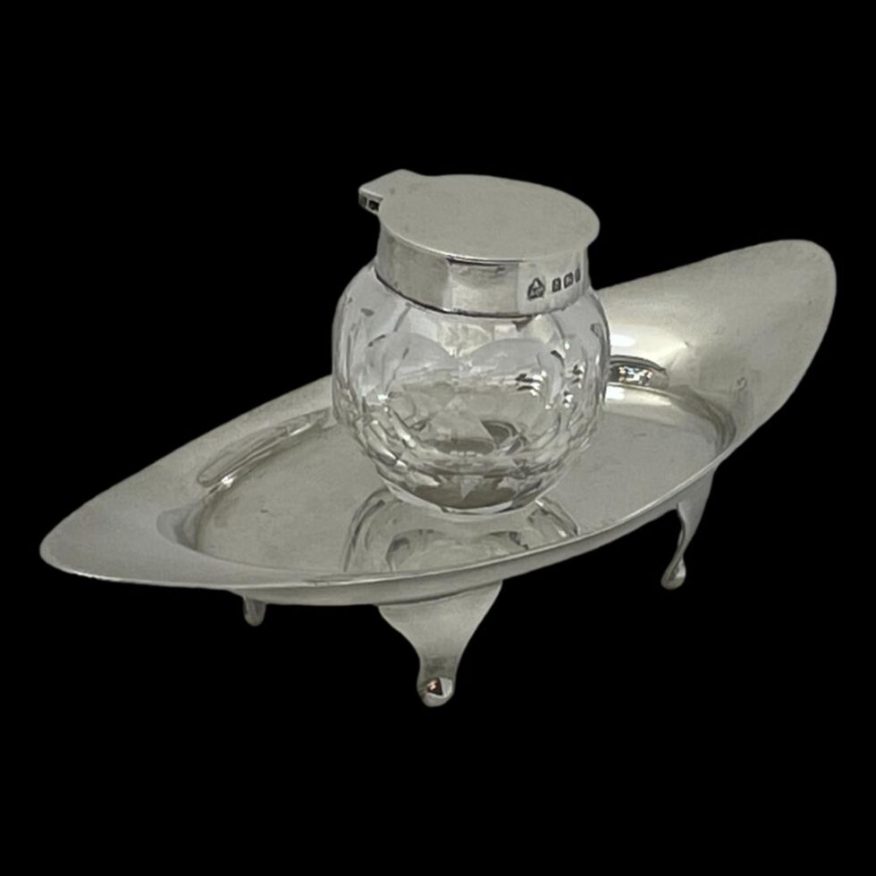 Photo of the Asprey silver and crystal inkwell and stand this time displayed from a side angle with the stand set diagonally across the page. The shot is taken from a slightly raised angle so the inside of the stand tray is visible.