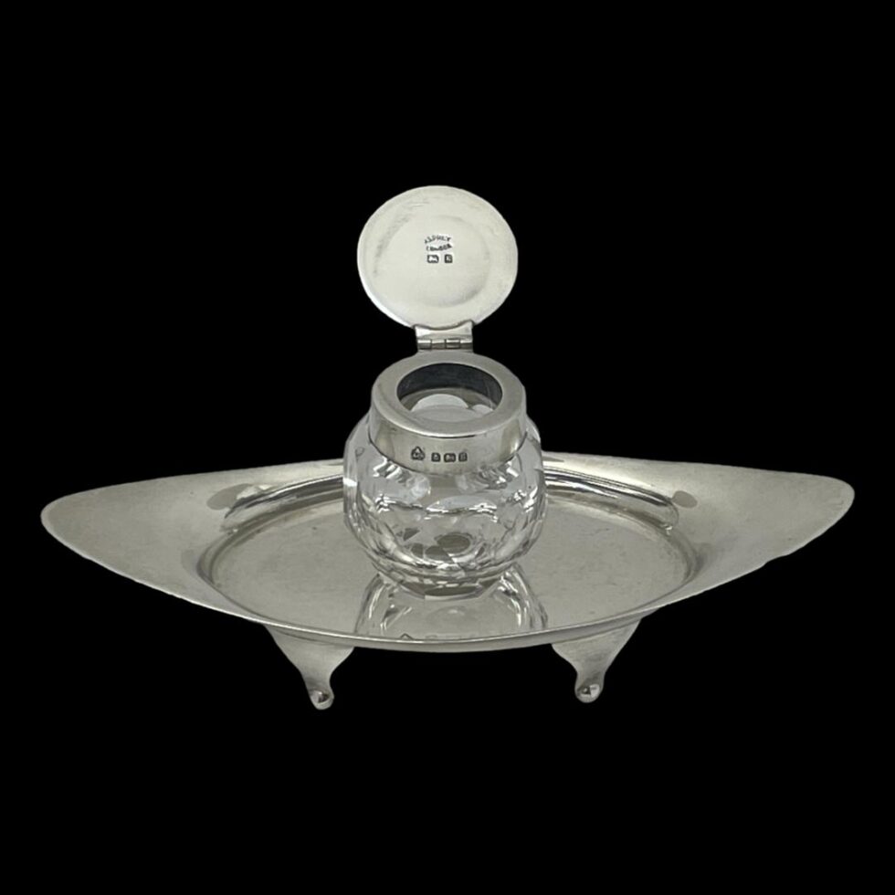 Photo looking down at the inkwell diagonally. The inkwell has its silver hinged lid open and hallmark on inside lid is visible as well as the hallmark to outer rim.