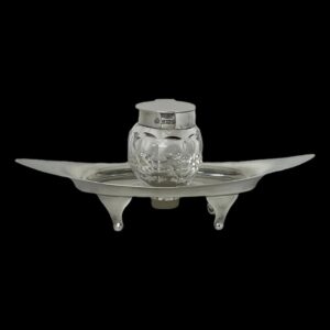 Main photo of Asprey silver &crystal inkwell seen from a near eye level. The silver base is eye shaped and stands on 4 feet. It has upward curved ends and a hole at the centre for the crystal inkwell to slot in to. The hallmark is visible around the outer rim if the inkwell.
