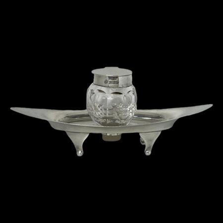 Main photo of Asprey silver &crystal inkwell seen from a near eye level. The silver base is eye shaped and stands on 4 feet. It has upward curved ends and a hole at the centre for the crystal inkwell to slot in to. The hallmark is visible around the outer rim if the inkwell.