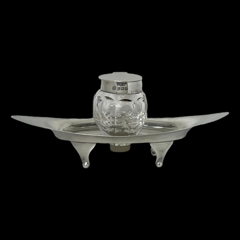 Main photo of Asprey silver &crystal inkwell seen from a near eye level. The silver base is eye shaped and stands on 4 feet. It has upward curved ends and a hole at the centre for the crystal inkwell to slot in to. The hallmark is visible around the outer rim if the inkwell.
