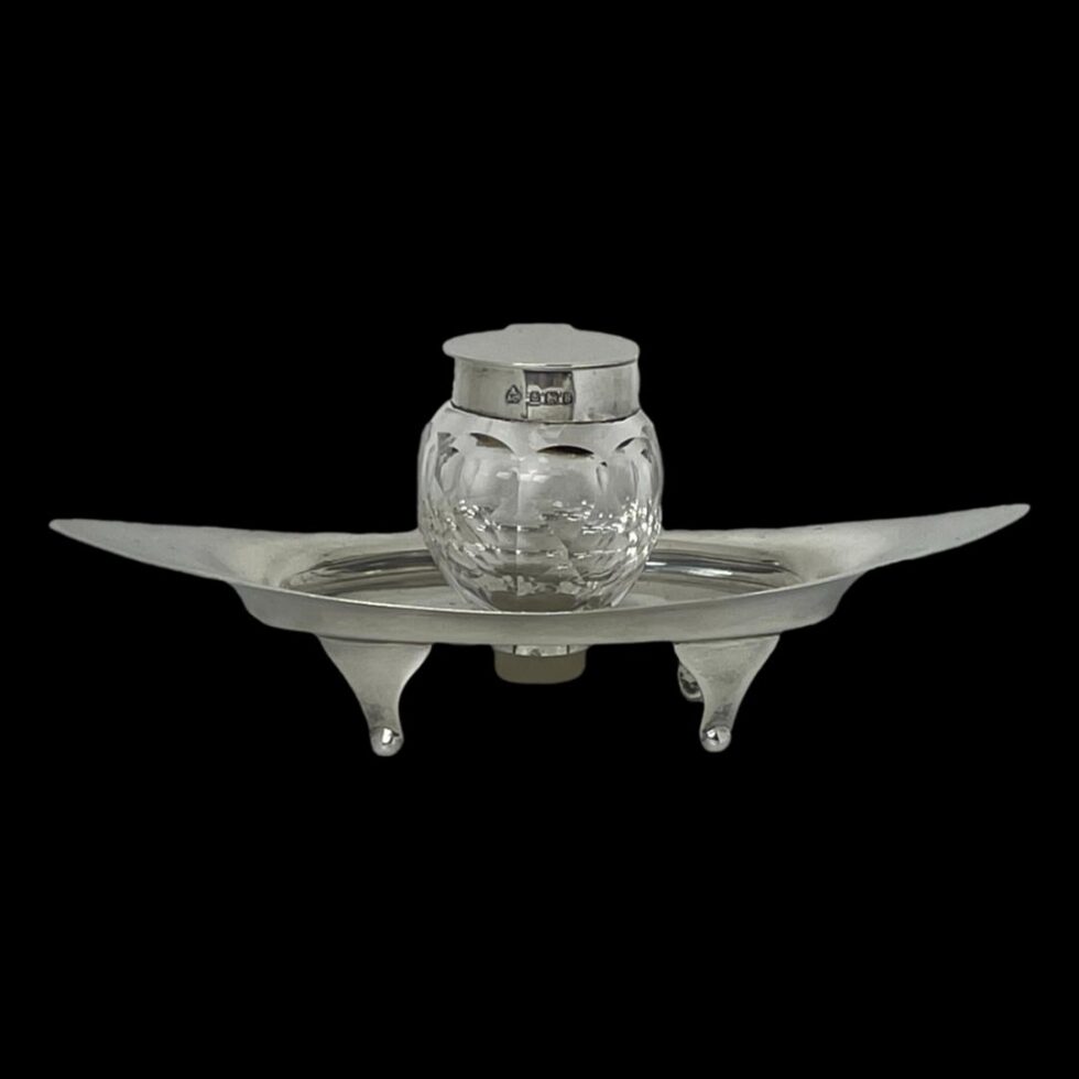 Main photo of Asprey silver &crystal inkwell seen from a near eye level. The silver base is eye shaped and stands on 4 feet. It has upward curved ends and a hole at the centre for the crystal inkwell to slot in to. The hallmark is visible around the outer rim if the inkwell.