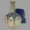 Main photo of Ada Tosen Doulton vase seen from an eye level angle. The base colour of the vase is a greyish green beige to the neck and cream to the bulbous bottom with an almost stained glass effect section in the middle with small blue and white flowers and green hearts. There are arches that decorate the upper area of the bulbous bottom which look like arched windows. A small rock of lapis stands to the back right of vase for effect.