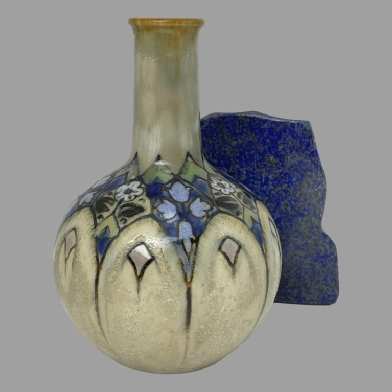 Main photo of Ada Tosen Doulton vase seen from an eye level angle. The base colour of the vase is a greyish green beige to the neck and cream to the bulbous bottom with an almost stained glass effect section in the middle with small blue and white flowers and green hearts. There are arches that decorate the upper area of the bulbous bottom which look like arched windows. A small rock of lapis stands to the back right of vase for effect.