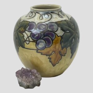 Main photo of Doulton Ethel Beard vase seen from an eye level. The base colour of the vase is a pale mustard on the lower end and a greyish cream at the top. The decoration on display in this photo is 2 dark green vine leaves partially visible on the left and right with a small bunch of purple grapes near centre of the leaves and a burnt orange colour leaf lower down right of grapes with lines of the vine visible in between and around the mouth. There is a small rock of natural amethyst crystal to the bottom left of vase which could be a small bunch of grapes with quite some stretch of the imagination!
