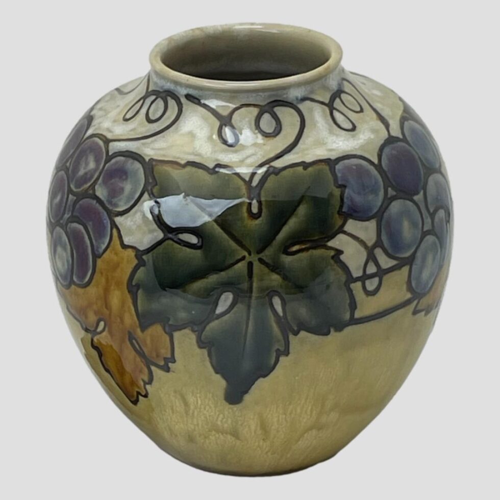 Photo of Ethel Beard Doulton vase seen a little in close up with a large dark green vine leaf in the centre foreground. The angle is slightly raised so peeking into the mouth a touch.