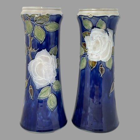 Main photo of Doulton tall rose vases seen side by side from an eye level. The vase on left is displaying the side with rose lower down and the one on right has rose at the top. The background colour of the vases is a rich royal blue with one large off white rose to each with a few rose buds in a brown colour and leaves in a mixture of green and light brown. The rim of the vases is a creamy white. The vases can be displayed like this (one rose higher, one lower) or with both showing roses at same height. The vases have a lovely chimney-like shape and are stunning even without flowers!
