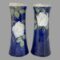 Main photo of Doulton tall rose vases seen side by side from an eye level. The vase on left is displaying the side with rose lower down and the one on right has rose at the top. The background colour of the vases is a rich royal blue with one large off white rose to each with a few rose buds in a brown colour and leaves in a mixture of green and light brown. The rim of the vases is a creamy white. The vases can be displayed like this (one rose higher, one lower) or with both showing roses at same height. The vases have a lovely chimney-like shape and are stunning even without flowers!