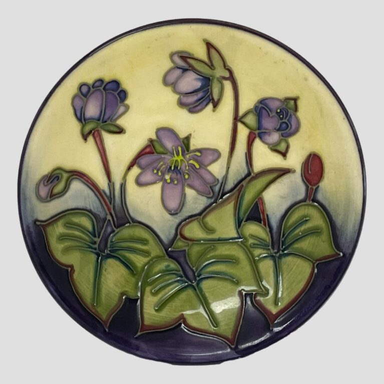 Main photo of Moorcroft Hepatica pin dish displayed upright with the inside of dish bowl on display. The base colours of the dish are cream 2 thirds at the top and purple a third at bottom. 4 lighter purple Hepatica flowers are in bloom with 2 buds one to left and other on right. The bud on right is a red colour like the stems of the flowers. To the bottom foreground are large green leaves red around the edges. A lovely gift item.