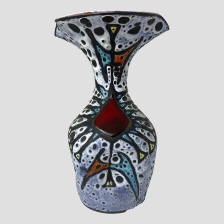Main photo of Vallauris fat lava vase seen from an eye level with the most decorated side on display. The photo is taken at eye level to show the form of the vase clearly. It is bottle shaped but with a large wide fluted top. The top of the vase is actually wider than the bottom. The base colour is a light blue with white and speckled black 'lava' gaps throughout. All the colours are matt except for the cherry red diamond shape just below the slender neck. The matt colours include red yellow and turquoise all within black lava beading.