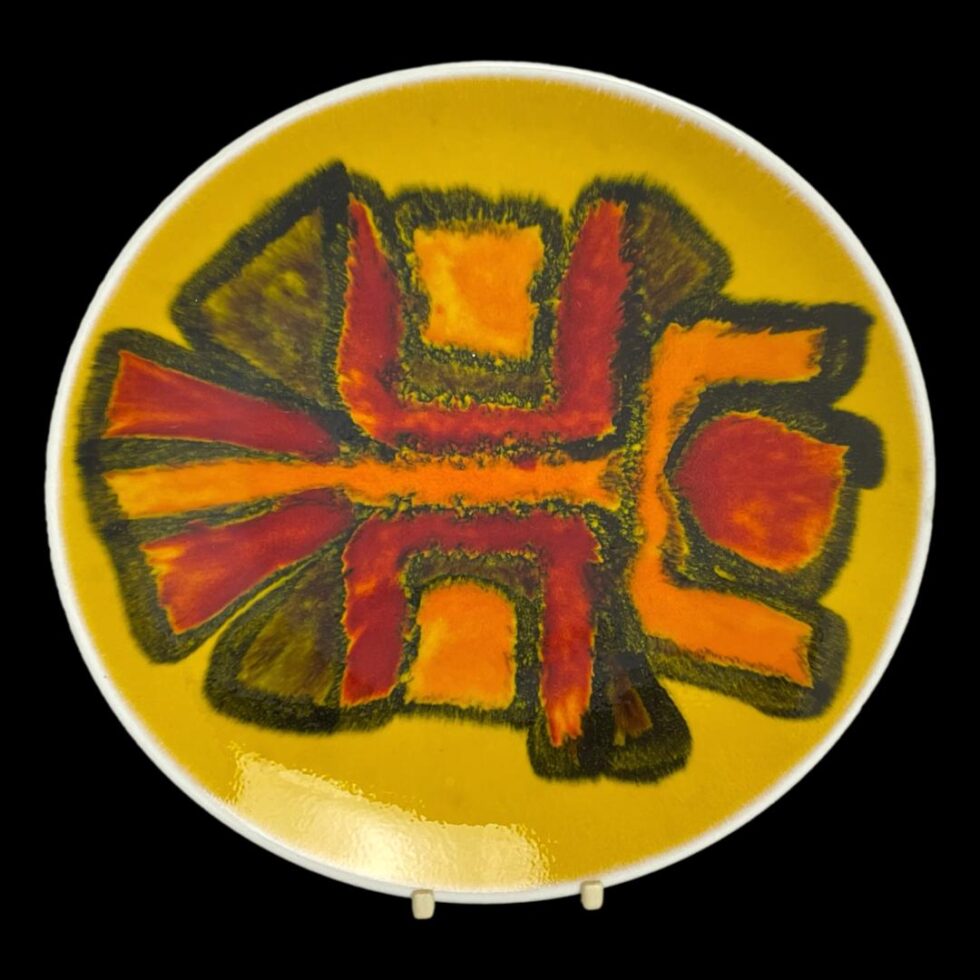 Main photo of Poole yellow Delphis dish displayed on a plate stand and against a black background. The abstract design to the centre in orange red and brown blocks of different shapes put together. To me looks like a person with arms raised in a dancing swirl.