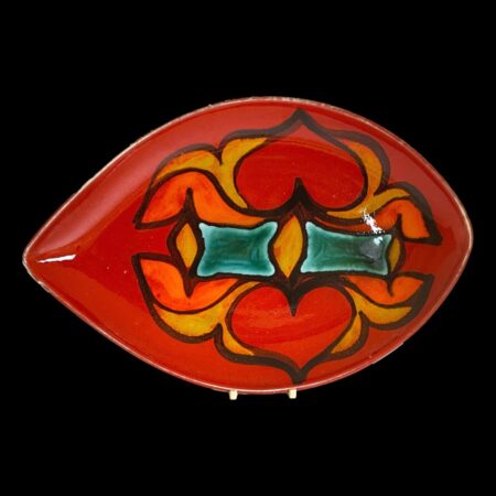 Main photo of red Poole Delphis dish displayed on a plate stand against a black background. The chilli red base colour is spicy hot with added fire from the yellow, orange & red design at the centre. The 2 rectangular blocks of turquoise added maybe to cool things down a touch? There is definitely something passion filled about this piece! The longer side of the almond shaped plate is on the left.