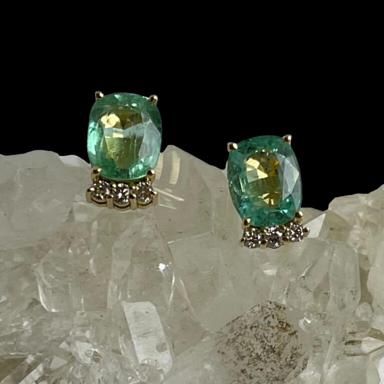 Main photo of gold emerald diamond studs displayed on top of a rock of crystal quartz with the earring fronts facing forward. The green colour of these emeralds is very unusual-more pale than regular emeralds. The diamonds are set below and slightly back from the emeralds and have intense sparkle.