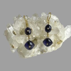 Main photo of gold iolite diamond earrings shown displayed hanging on a rock of natural quartz crystal with the earring fronts facing forward. The earrings construction begins with an 18 karat gold hook fastener. An oval iolite cabochon with a small round cut diamond to the centre is first followed below it by a larger round cut diamond divider. At the bottom of the earring is a square iolite cabochon set with one corner at top like diamond shape and there is another small round cut diamond to the centre of this. The iolite is a beautiful darker blue with tinges of purple in certain light. They marry beautifully with the colour of the 18 karat gold.