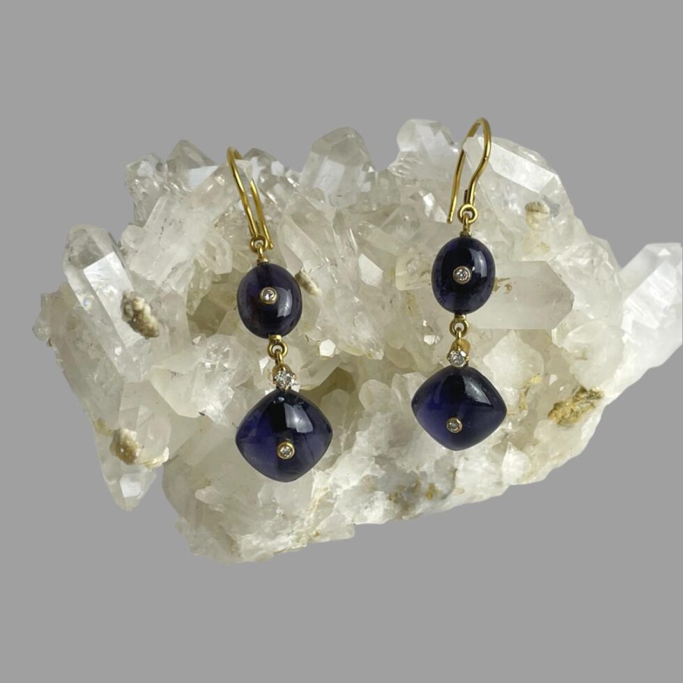 Main photo of gold iolite diamond earrings shown displayed hanging on a rock of natural quartz crystal with the earring fronts facing forward. The earrings construction begins with an 18 karat gold hook fastener. An oval iolite cabochon with a small round cut diamond to the centre is first followed below it by a larger round cut diamond divider. At the bottom of the earring is a square iolite cabochon set with one corner at top like diamond shape and there is another small round cut diamond to the centre of this. The iolite is a beautiful darker blue with tinges of purple in certain light. They marry beautifully with the colour of the 18 karat gold.